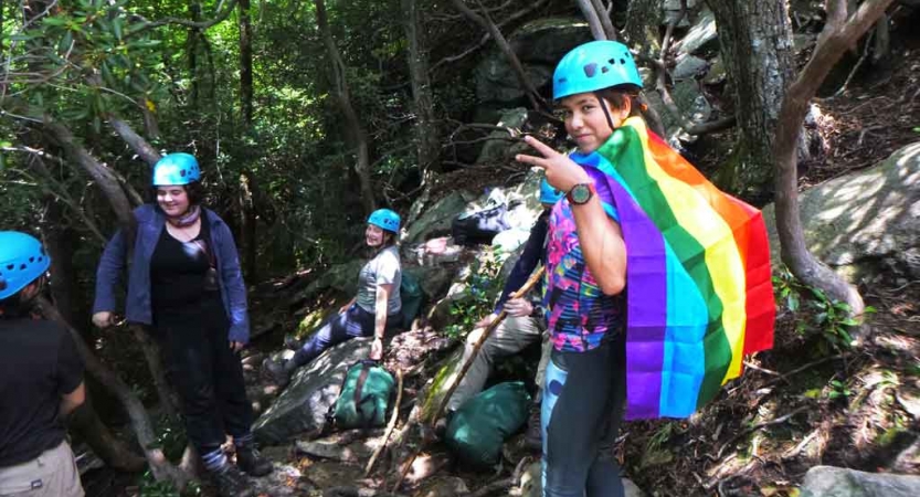 a student wearing a rainbow flag as a cape gives a peace sign while on a rock climbing trip in north carolina